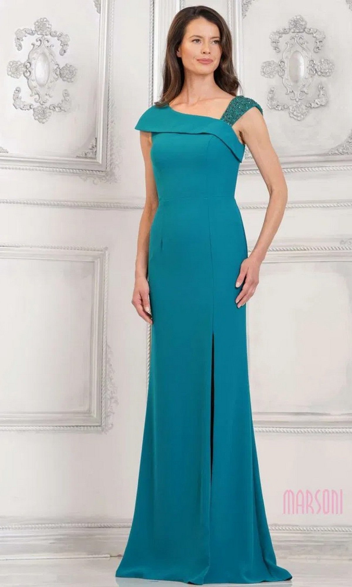 A woman in a teal green Marsoni MV1296 long formal dress with an off-the-shoulder neckline and front slit stands against an ornate white wall.